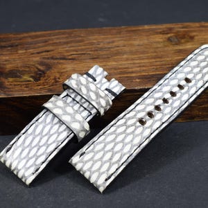 Può includere: Cinturino per orologio in due pezzi con motivo a pelle di serpente grigio e bianco. Il cinturino è in pelle e presenta cuciture nere. Il cinturino è su un blocco di legno.