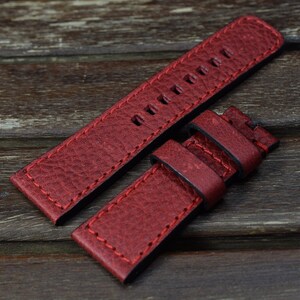 May include: Red leather watch strap with black stitching and a buckle. The strap is made of a textured leather and has a classic design.