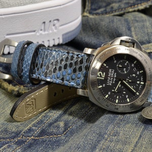 May include: A black and silver chronograph wristwatch with a blue and black patterned leather strap. The watch face has white markings and the text "LUM NOR PANERAI Day ight" printed on it.