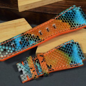 Peut inclure: Deux bracelets de montre avec un motif de peau de serpent coloré. Les bracelets présentent un mélange vibrant de teintes bleues, oranges et noires, avec des bords orange vif. Les bracelets sont en cuir et sont conçus pour une montre.