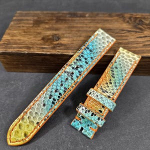 以下が含まれることがあります： 青、黄、黒の色合いのヘビ革模様の2ピースの時計ストラップ。オレンジ色のステッチとエッジが付いています。時計バンドは革製です。