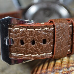 May include: A brown leather watch strap with white stitching and a silver buckle. The strap has a textured surface and is designed for a wristwatch.