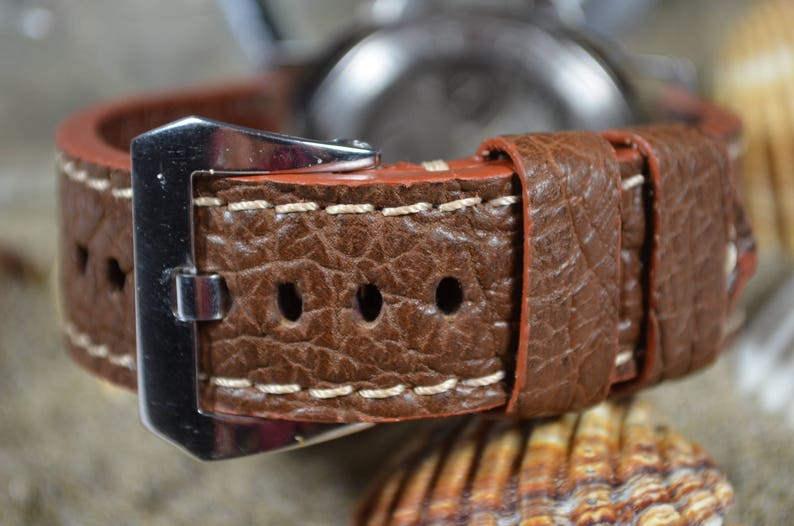 May include: A brown leather watch strap with white stitching and a silver buckle. The strap has a textured surface and is designed for a wristwatch.