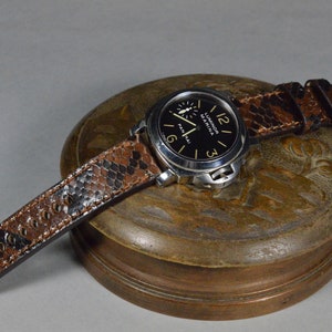 Può includere: Un orologio nero e argento con un cinturino in pelle marrone. Il quadrante dell'orologio ha uno sfondo nero con segni bianchi e le parole "Luminor Marina" e "Panerai".