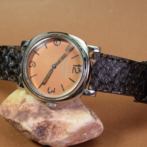 Op de afbeelding: Een zilveren polshorloge met een bruin leren bandje. De wijzerplaat van het horloge is oranje met zwarte Romeinse cijfers voor de uren en de tekst "RADIOMIR PANERAI" erop gedrukt.
