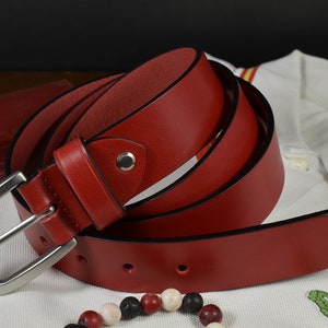 Può includere: Una cintura in pelle rossa con una fibbia argentata. La cintura è piegata e appoggiata su una superficie bianca.