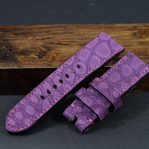 Può includere: Un cinturino viola con un motivo strutturato a forma di coccodrillo. Il cinturino ha cuciture rosa e diversi fori per la regolazione. Il cinturino è realizzato in un materiale resistente.