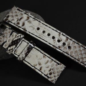 Pode incluir: Uma pulseira de relógio de couro branco com um padrão de pele de cobra. A pulseira tem uma fivela e costuras ao redor das bordas.