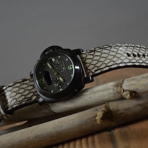 Puede incluir: Un reloj negro con una correa de cuero blanca con un patrón de piel de serpiente. El reloj está sobre una superficie de madera.