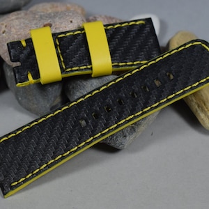 Op de afbeelding: Zwart horlogeband met gele stiksels en een carbonfiber patroon. Het band is gemaakt van leer en heeft een gespsluiting.