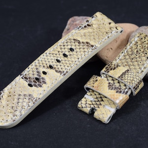 May include: Two-piece watch strap set with a snakeskin pattern in beige, brown, and black. The watch straps are made of leather and are displayed on a dark surface with rocks in the background.