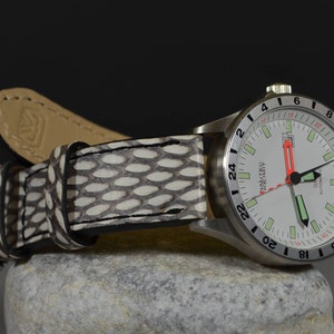 May include: A silver wristwatch with a white face and black markings. The watch has a white leather strap with a black and white pattern. The watch face has a 24-hour display and the words "Nautec" and "Automatic" printed on it.