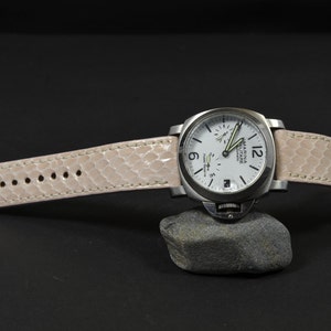 May include: A stainless steel wristwatch with a white face and a light pink snakeskin-patterned band. The watch is resting on a small gray rock against a black background. The watch face has black numerals and hands.
