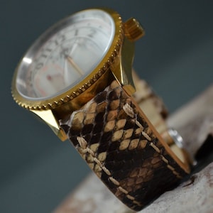 Pode incluir: Um relógio de pulso dourado com um mostrador branco e uma pulseira de pele de cobra castanho e bege.