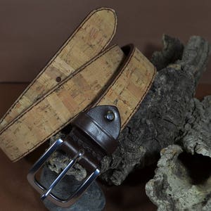 May include: A brown leather belt with a cork buckle. The belt is wrapped around a piece of driftwood.