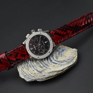 May include: A silver chronograph wristwatch with a black face and red snake skin band. The watch is resting on a gray and white textured surface.