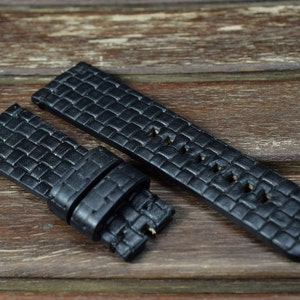 Puede incluir: Correa de reloj de cuero negro con un patrón cuadrado y agujeros para ventilación. La correa se muestra sobre una superficie de madera.
