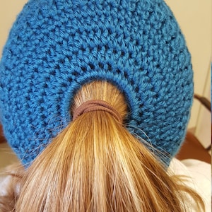 Puede incluir: Un gorro azul de ganchillo con un agujero en la parte posterior para una coleta.