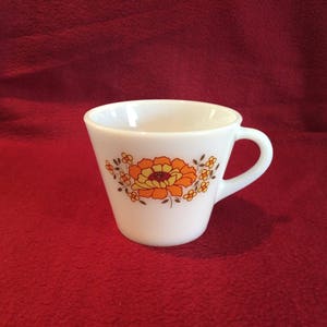 May include: White ceramic teacup with a floral design in orange and yellow. The cup has a handle and a flared shape. The floral pattern is centered on the cup and includes small flower accents.