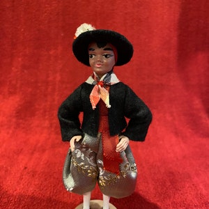May include: A small, black-haired doll wearing a black jacket, red sash, and patterned pants. The doll is wearing a black hat with a white pom-pom and is standing on a beige base.