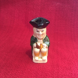 Może przedstawiać: Ceramiczna figurka mężczyzny w czarnym kapeluszu i zielonej kurtce, trzymającego kubek. Figurka siedzi na białej podstawie.