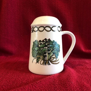 Puede incluir: Salero de cerámica blanca con asa y tapa. El salero presenta un diseño floral con flores azules y verdes atadas con una cinta amarilla. Un diseño de cuerda negra y amarilla decora la parte superior.