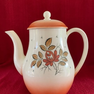Könnte beinhalten: Eine weiße Teekanne aus Keramik mit einem Blumendesign. Die Teekanne hat eine rote Rose mit braunen Blättern und einen weißen Deckel mit orangefarbener Oberseite.