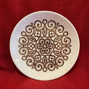 May include: A white ceramic plate with a brown and white geometric design. The design features a repeating pattern of swirls and flowers.
