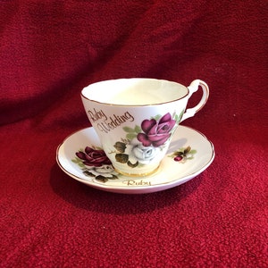 May include: A white teacup and saucer with a gold rim. The teacup features a floral design with red and white roses. The saucer has the words "Ruby Wedding" and "Ruby" printed on it.