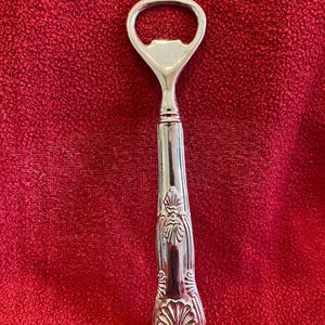 May include: A silver bottle opener with an ornate handle. The opener has a loop at the top for leverage and a detailed design on the handle. The handle features a floral pattern. The opener is made of metal.