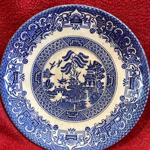 May include: A round, blue and white ceramic plate with a classic Willow pattern design. The plate features a central scene of a pagoda, bridge, and trees, surrounded by a decorative border. The plate is set against a red background.