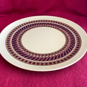 Könnte beinhalten: Ein weißer Keramik-Dinnerteller mit einem braunen und goldenen geometrischen Design. Das Design zeigt eine Reihe konzentrischer Kreise mit einem sich wiederholenden Muster aus Quadraten und Zickzacklinien.