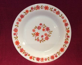 May include: A white ceramic dinner plate with a floral pattern in shades of orange and brown. The plate has a scalloped edge and a raised rim.