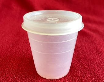 Puede incluir: Un vaso de plástico transparente con tapa blanca. El vaso tiene forma cilíndrica con estrías horizontales y un borde ligeramente ensanchado. La tapa es plana con un borde elevado.