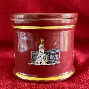 Puede incluir: Un tarro de cerámica burdeos con un borde dorado y una imagen blanca del Palacio de Buckingham y el Victoria Memorial. El tarro tiene una tapa.