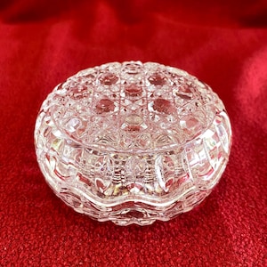 May include: A clear, round crystal trinket box with a patterned lid. The box is made of cut glass, with a detailed geometric design. The box sits on a red surface, and the light reflects off the facets of the crystal.