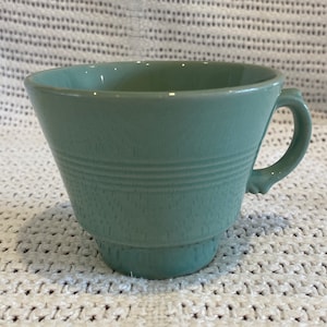 May include: A pale green ceramic teacup with a wide, flared rim and a handle. The cup has a textured surface and three horizontal lines around the middle.