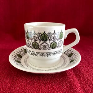 May include: White teacup and saucer set with a repeating pattern of green and black stylized leaf shapes. The cup has a curved handle and sits on a matching saucer. The set is on a red background.