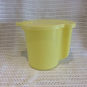 Può includere: Un'ampolla di plastica gialla con un coperchio. L'ampolla ha un manico curvo e un'ampia apertura.