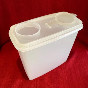 May include: A clear, rectangular food storage container with a white lid. The lid has a circular opening and a small, round, hinged lid. The container is made of translucent plastic and sits on a red surface.