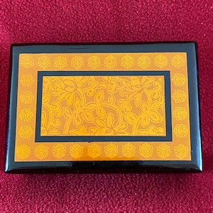 Op de afbeelding: Een zwarte houten doos met een gouden bloemenontwerp. De doos heeft een scharnierend deksel en een decoratieve rand.