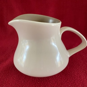 Puede incluir: Una jarra de crema de cerámica blanca con un borde marrón y un asa curva. La jarra tiene una forma redondeada y bulbosa.