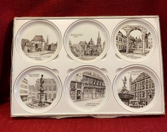 May include: A set of six white ceramic coasters, each featuring a black and white illustration of a building or landmark from Bad Aachen, Germany. The coasters are arranged in a white cardboard box.