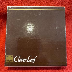 May include: A brown box with the words "Clover Leaf" in white script. The box has a gold border and a logo in the lower left corner. The background is a red, textured surface.