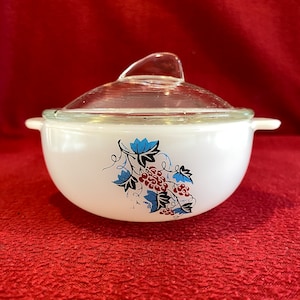 Puede incluir: Cazuela blanca con tapa de cristal transparente. El plato presenta un diseño floral con detalles azules y rojos. El plato tiene dos asas pequeñas a cada lado.