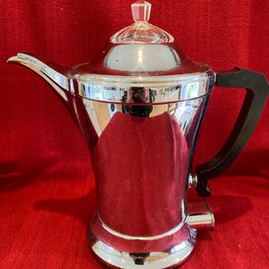 May include: A vintage chrome coffee pot with a black handle and a clear glass lid. The pot has a polished, reflective surface and a spout for pouring. The design is classic and elegant, suitable for serving coffee or tea.