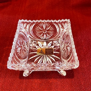 Può includere: Piatto quadrato di cristallo trasparente con un motivo floreale e tre piedini. Il piatto ha un bordo smerlato e un'etichetta al centro. Il piatto di cristallo è probabilmente per uso decorativo.