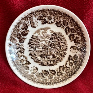 May include: A white and brown decorative plate with a detailed landscape scene in the center. The plate features a floral and leaf border design. The landscape scene depicts a building, trees, and animals. The plate is set against a red background.