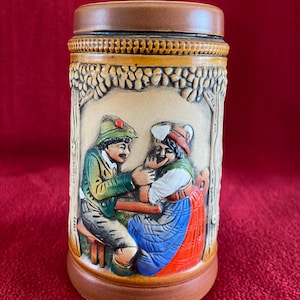 Peut inclure: Une chope à bière en céramique avec un extérieur brun et beige. La chope présente un motif en relief d'un homme et d'une femme en costume traditionnel allemand. L'homme porte une veste verte et un pantalon marron, tandis que la femme porte une robe bleue et rouge. La chope a un couvercle brun.