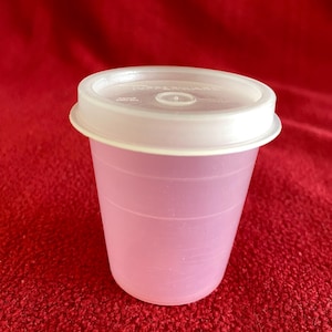 Puede incluir: Un vaso de plástico rosa translúcido con tapa blanca. El vaso tiene forma cilíndrica con estrías horizontales. La tapa es plana con un borde ligeramente elevado. El fondo es de color rojo.